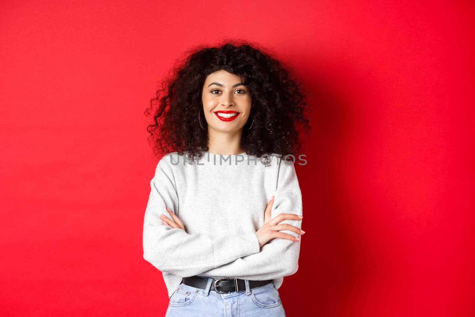 Confident smiling woman with makeup and curly hairstyle, cross arms on chest and looking professional, red background.