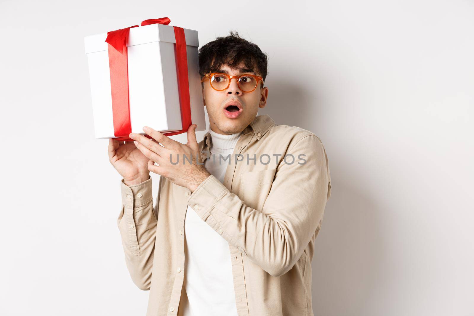 Holidays concept. Funny young man in glasses shaking gift box, listening what inside present and gasping amazed, standing on white background.