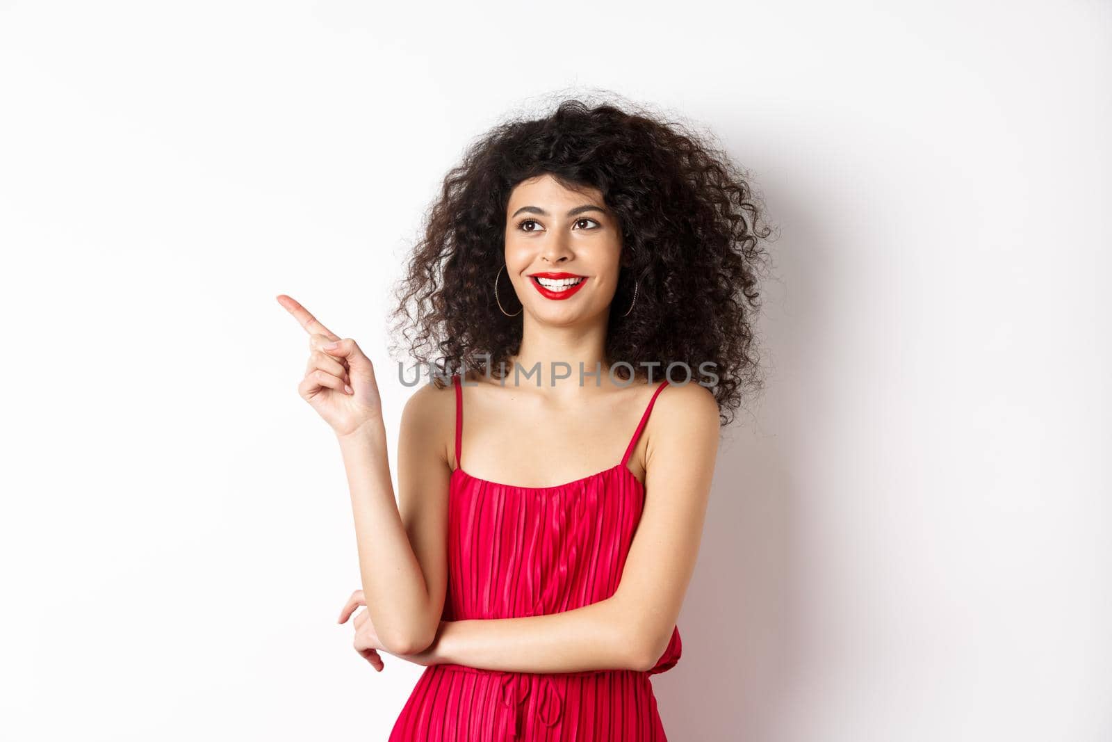 Elegant girl in red dress and evening makeup, pointing finger left and looking at logo, smiling satisfied, standing on white background by Benzoix