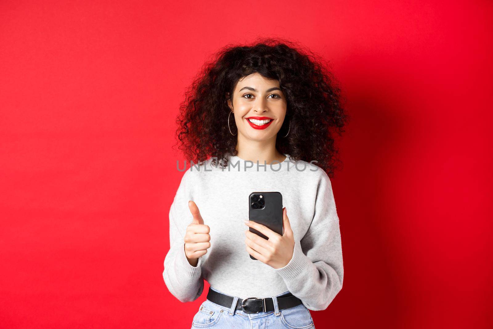 Smiling european woman looking happy and satisfied, using mobile phone and making thumb up, recommending application, standing on red background.