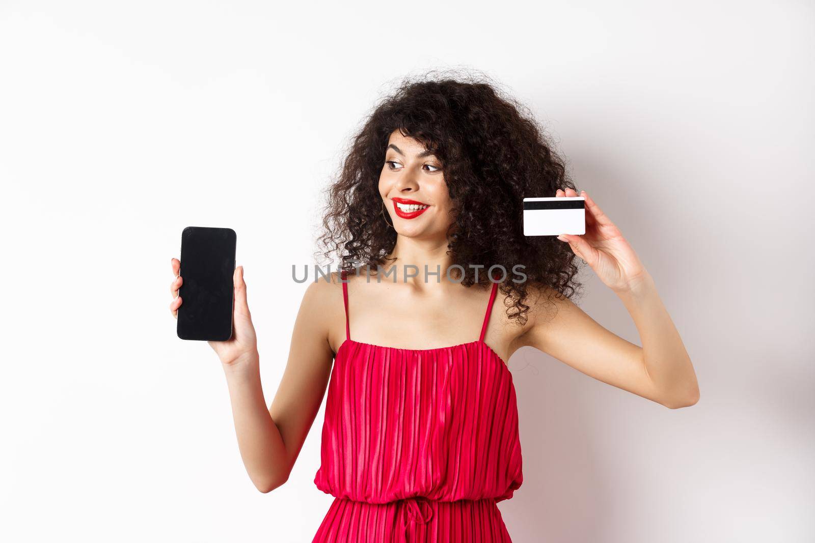 Online shopping concept. Elegant curly-haired woman in red dress showing plastic credit card and empty mobile phone screen, standing over white background by Benzoix