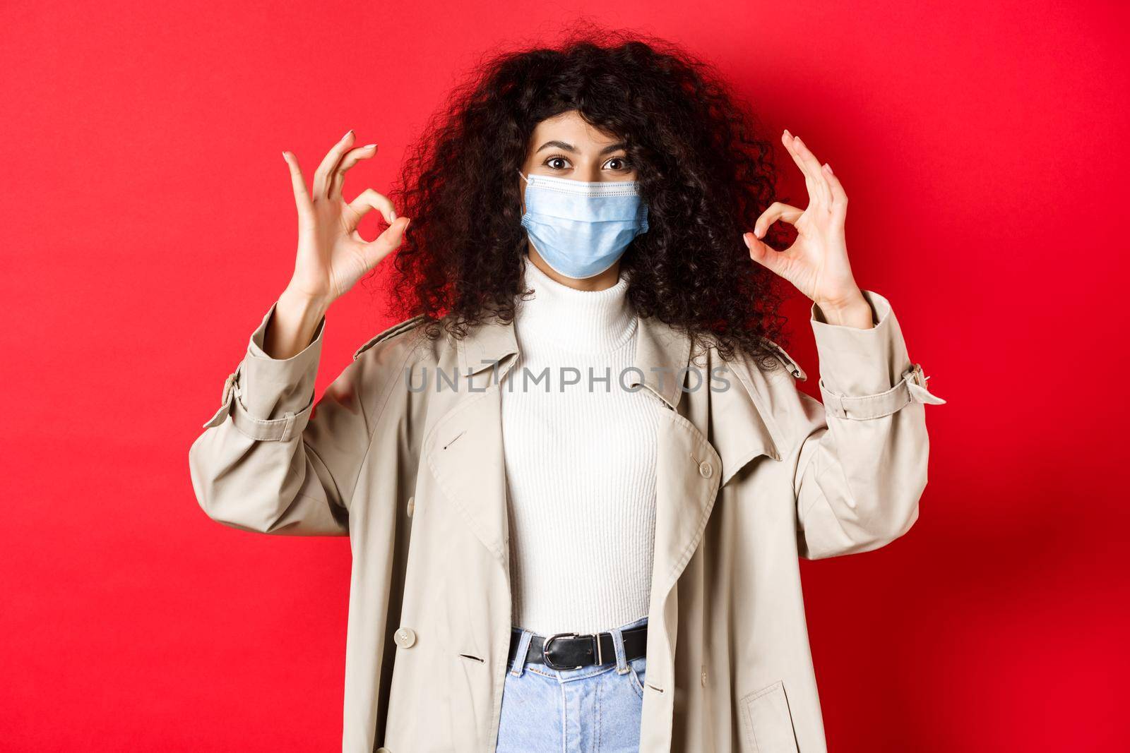 Covid-19, social distancing and quarantine concept. Fashionable woman with curly hair, wearing medical mask and trench coat, showing okay gestures, red background.