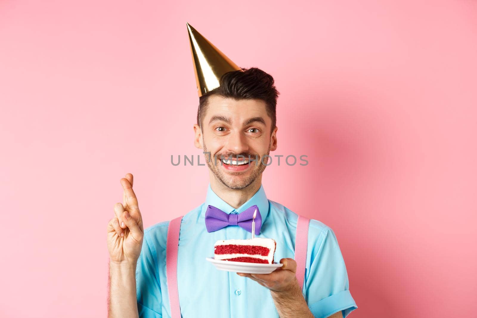 Holidays and celebration concept. Happy young man enjoying birthday party, wearing cone hat and cross fingers, making wish on bday cake with candle, pink background by Benzoix