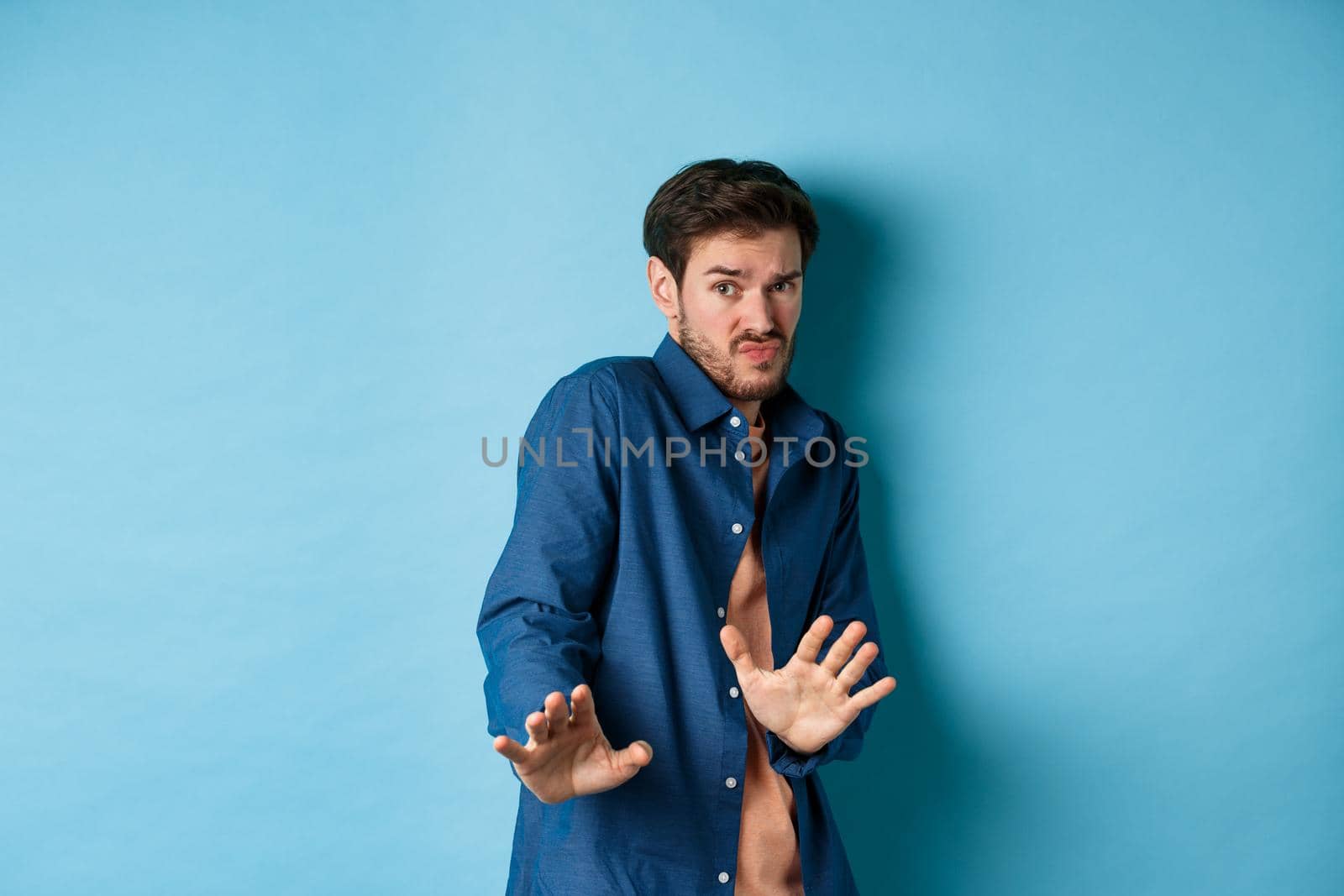 Timid young man asking to keep away, stretch out hands defensive and step back, refusing something disgusting, standing on blue background by Benzoix
