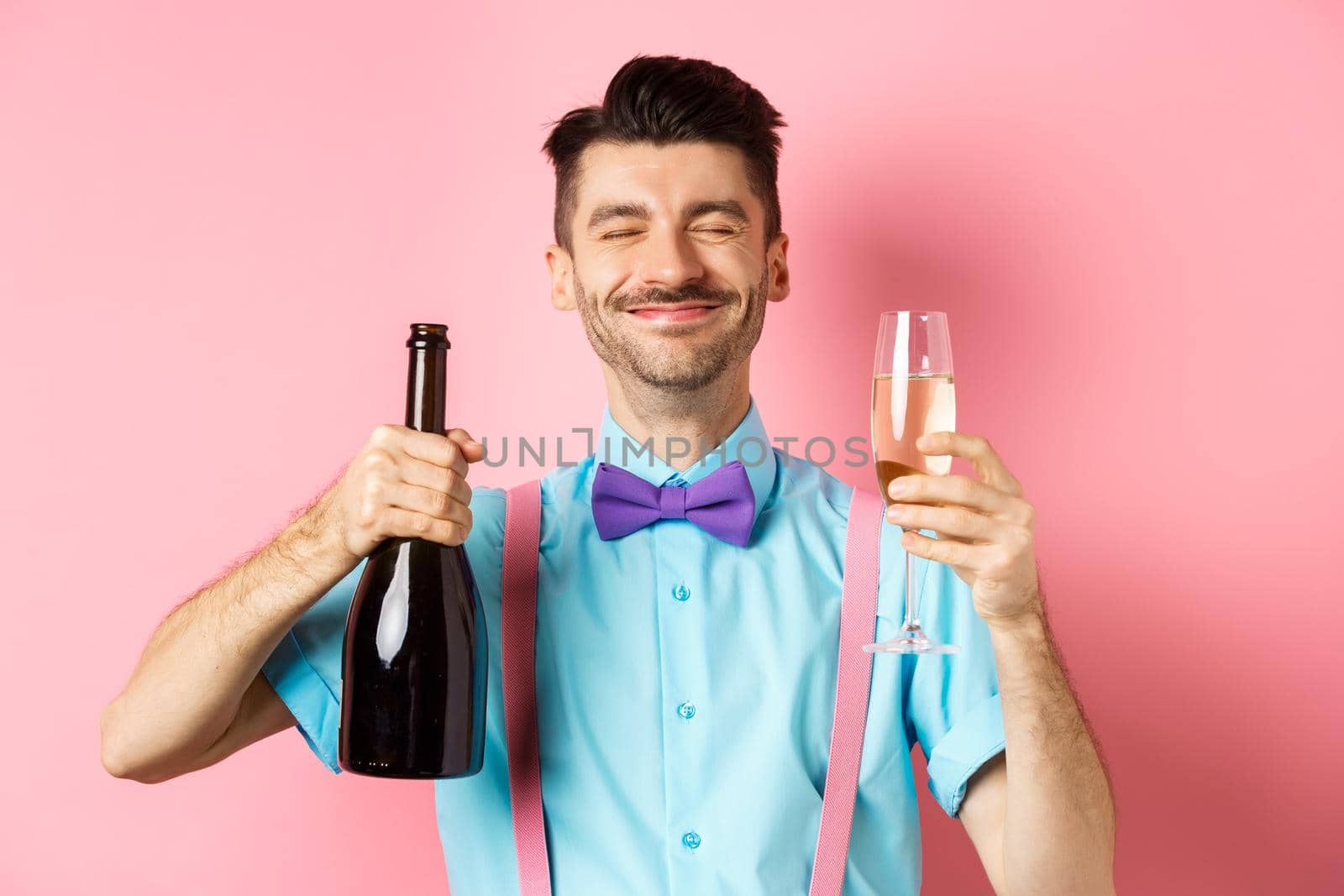 Holidays and celebration concept. Happy man smiling and enjoying drinking at party, holding bottle of champagne and glass, standing over pink background.