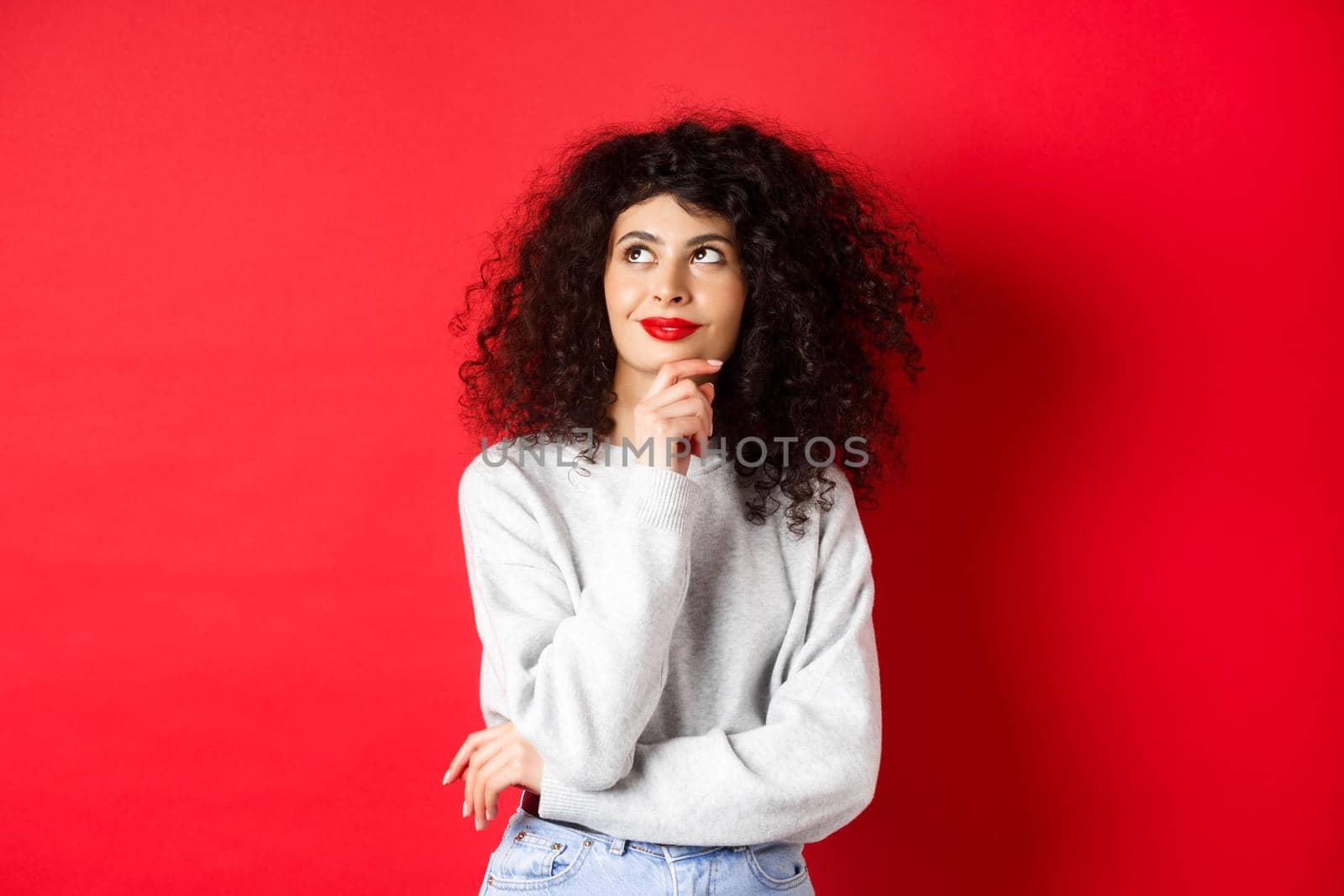 Pensive stylish woman with curly hair, red lips, looking thoughtful at upper left corner logo and smiling, standing against studio background by Benzoix