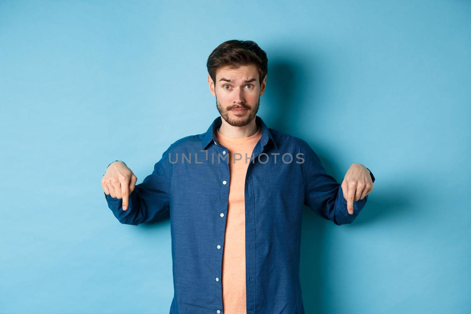 Confused male model in casual clothes, looking puzzled and pointing fingers down, showing banner, standing on blue background by Benzoix