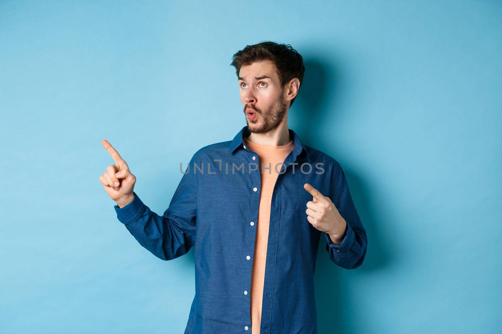 Impressed guy looking and pointing at upper left corner, showing advertisement, standing on blue background.