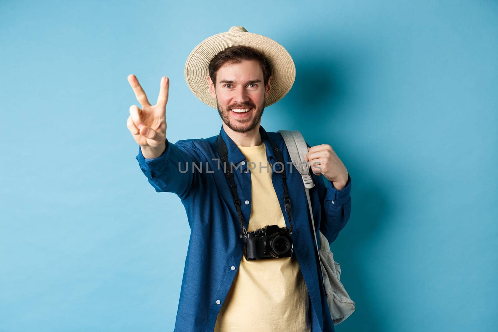 Handsome positive guy tourist showing peace sign on summer vacation, enjoying tropical holidays under sun, wearing straw hat, holding backpack and camera, blue background.