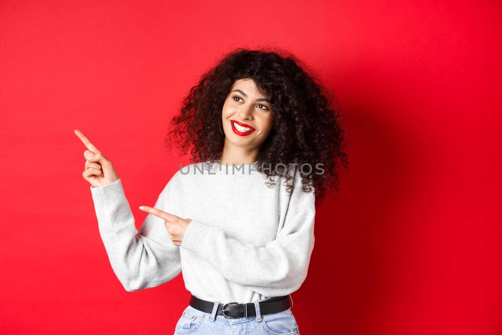 Beautiful european woman with curly hair and makeup, pointing fingers left and looking aside with dreamy smile, checking out promotion deal, red background.