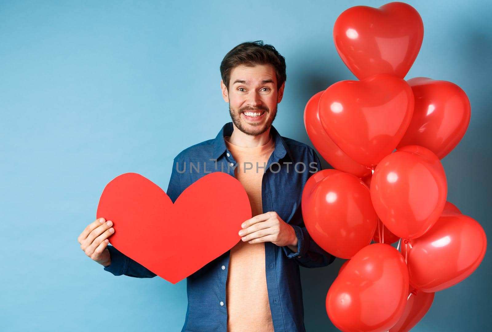 Valentines day concept. Smiling man say I love you, holding paper red heart cutout, standing near romantic balloons, blue background.