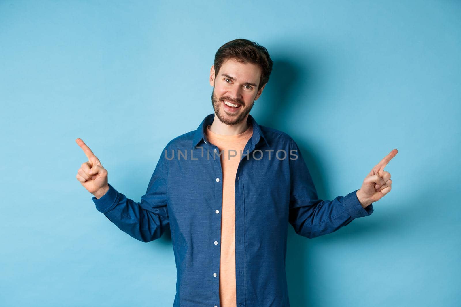 Handsome caucasian man with beard, smiling with white teeth and pointing fingers sideways, showing two choices, standing on blue background.