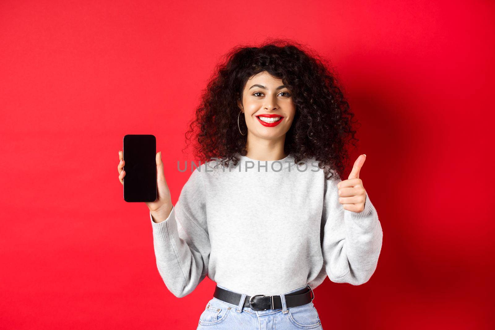 Portrait of attractive smiling woman with curly hair, showing empty mobile phone screen and thumb-up, recommending online promo, standing on red background.