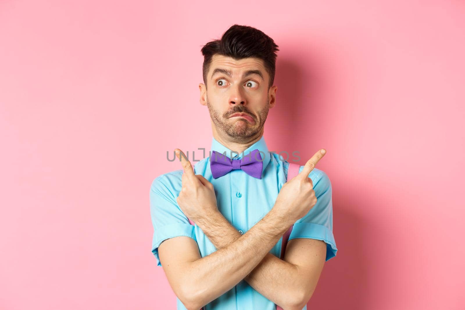 Puzzled funny man in bow-tie pointing fingers sideways, showing two variants on both sides, looking confused, standing over pink background.