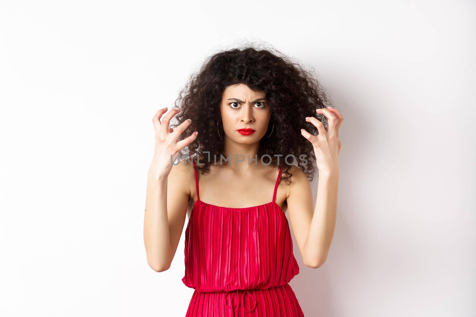 Angry italian woman with curly hair and red dress, boiling from anger, frowning and clenching hands outraged, standing on white background.