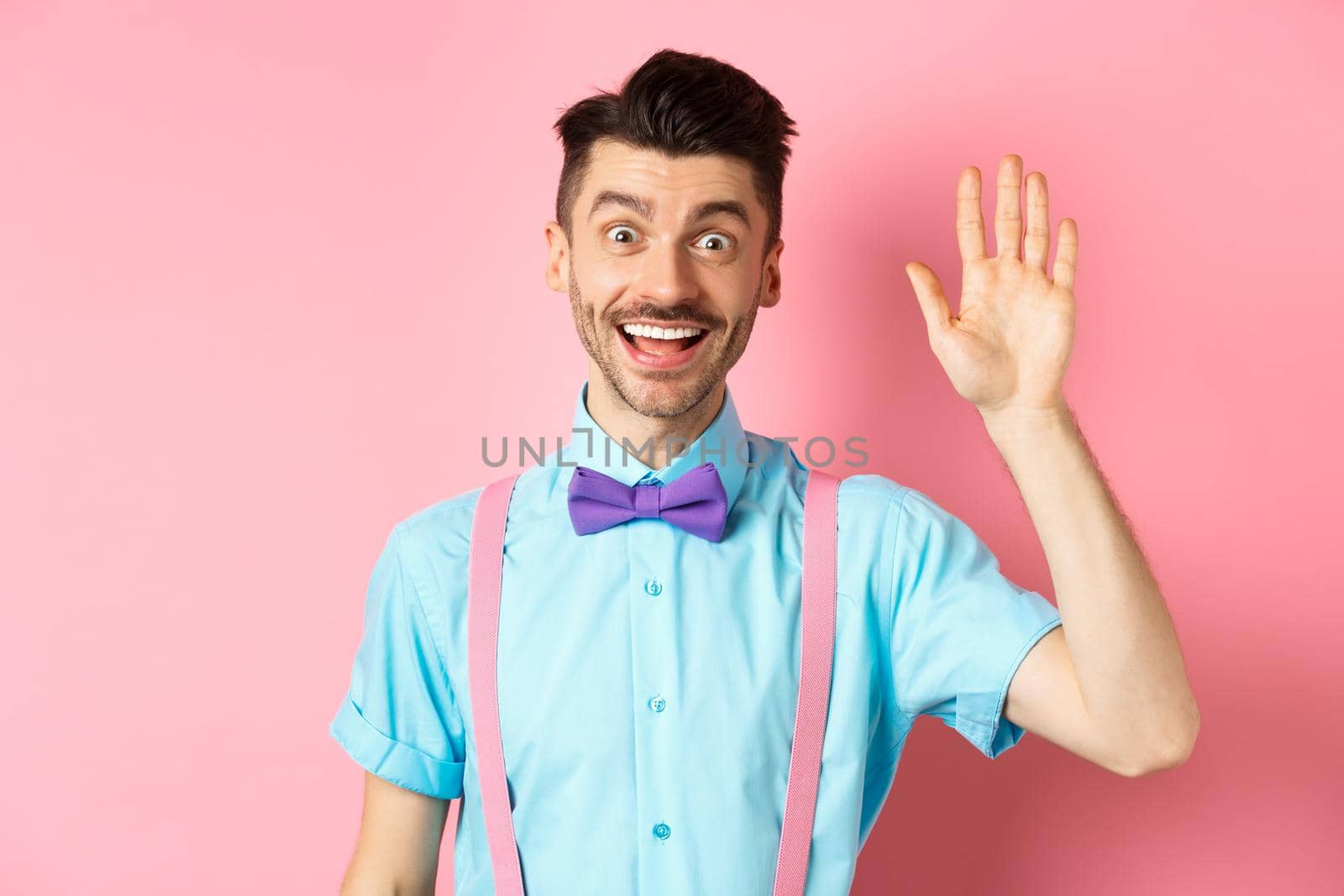 Friendly smiling man in funny bow-tie saying hello, waving hand to greet you, make hi gesture and looking happy yo see you, standing over pink background.