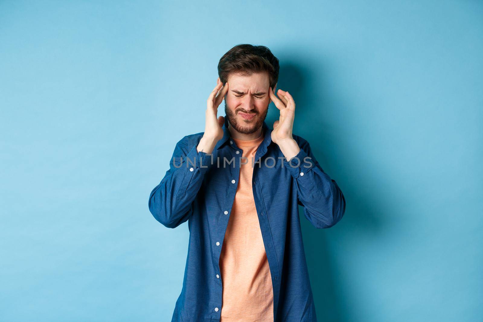 Man feeling sick, touching head and grimacing from painful migraine, having headache or feel dizzy, standing on blue background by Benzoix