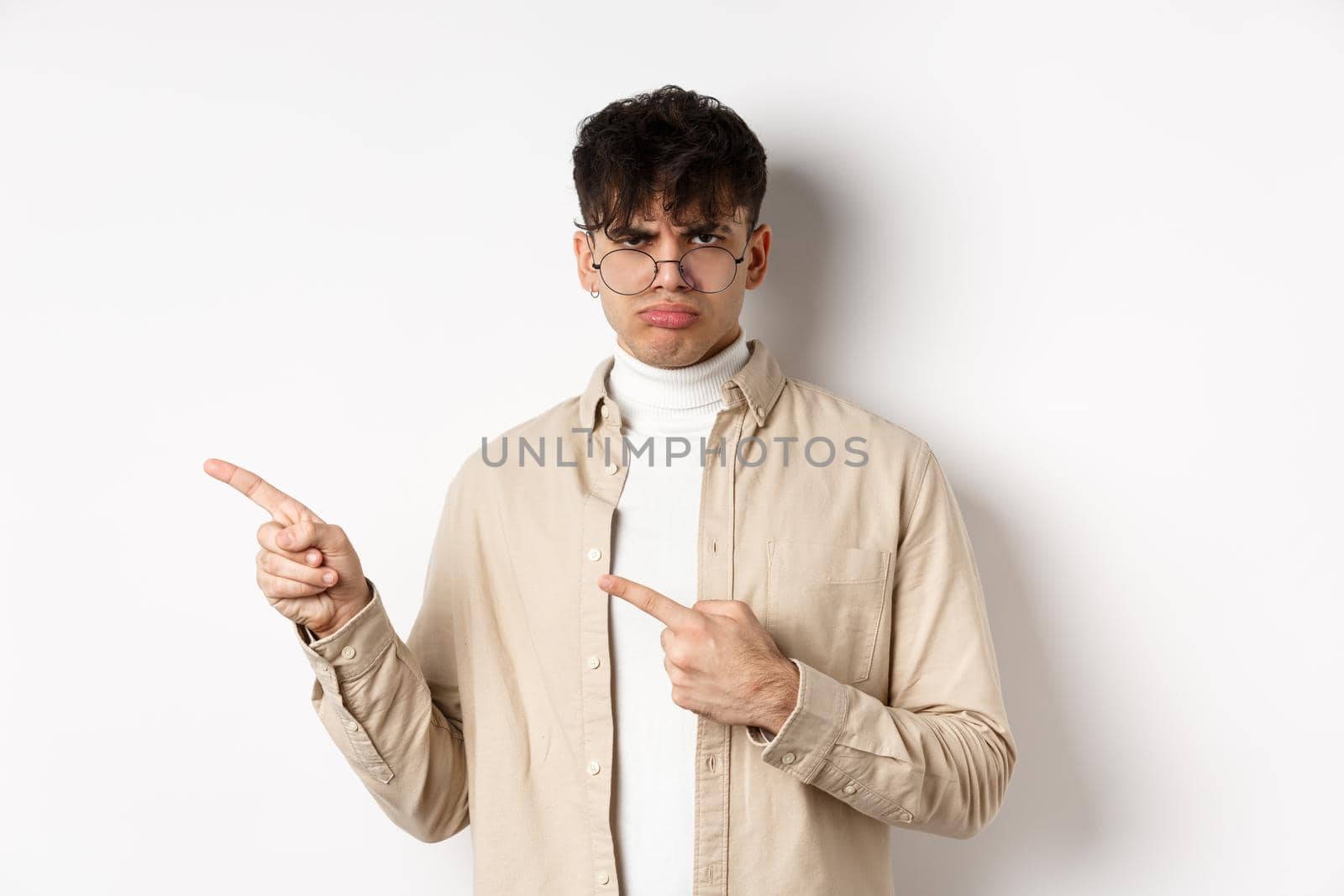 Sad and grumpy boy pointing fingers left, sulking with childish expression, complaining on something bad, standing on white background by Benzoix