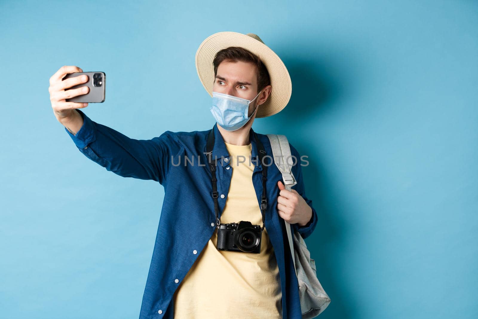 Covid-19, pandemic and travel concept. Tourist on summer vacation taking selfie in medical mask, photographing on smartphone, standing on blue background.