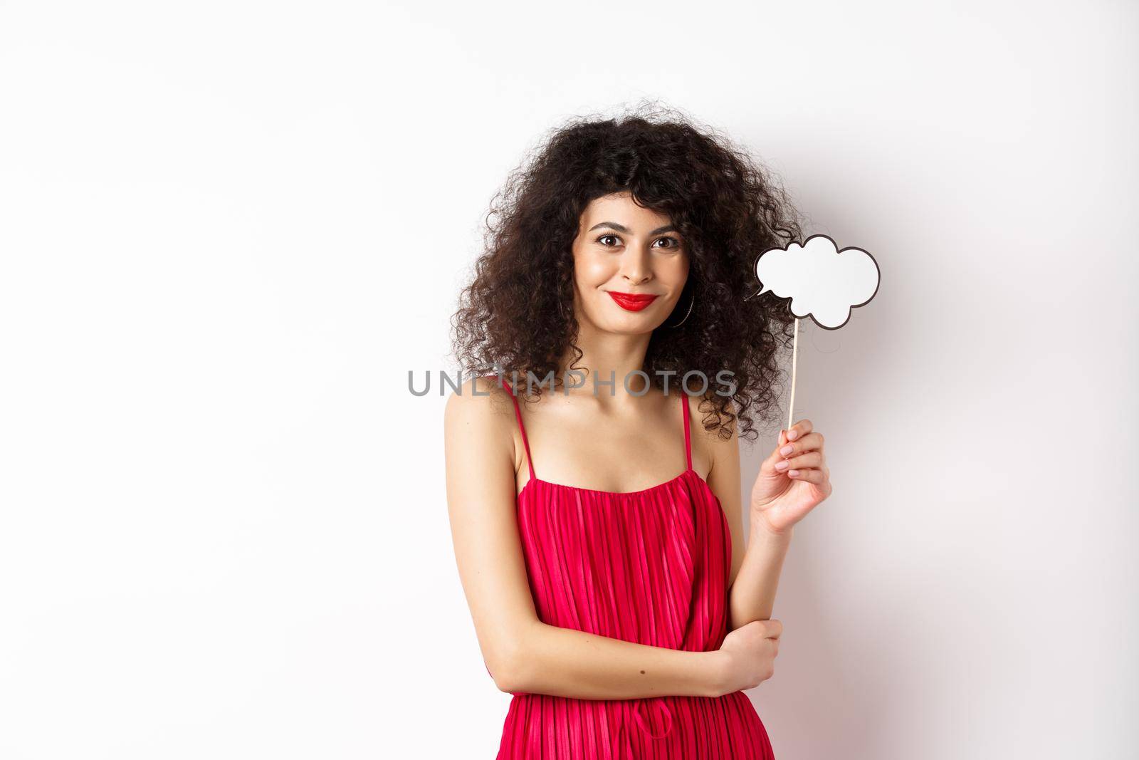 Elegant woman with curly hair, wearing red evening dress, holding cloud and smiling, standing on white background.