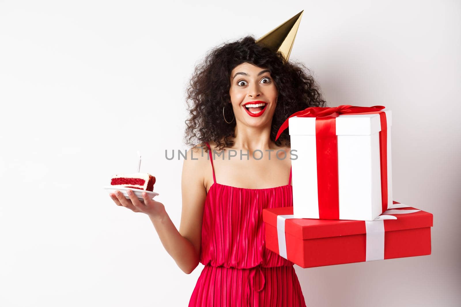 Cheerful girl in party hat and red dress, celebrating birthday, holding surprise gift and b-day cake, enjoying holiday, standing over white background.