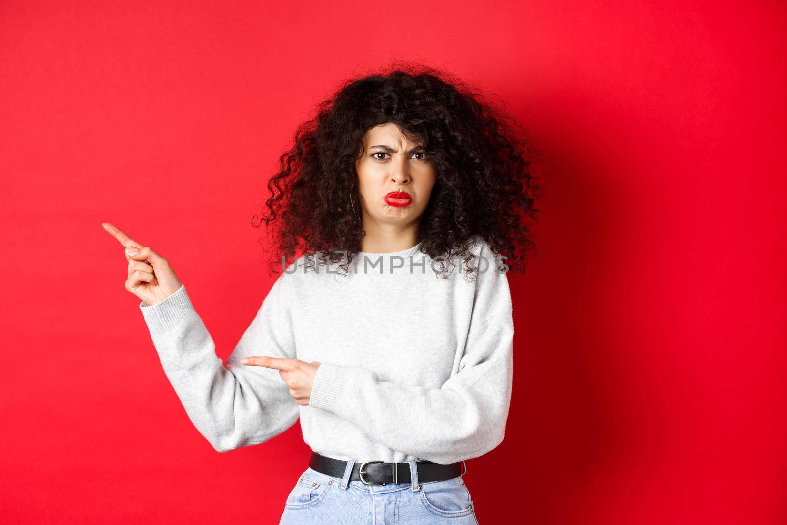 Disappointed funny woman, frowning and complaining, pointing fingers left at empty space and arguing, looking displeased, red background.