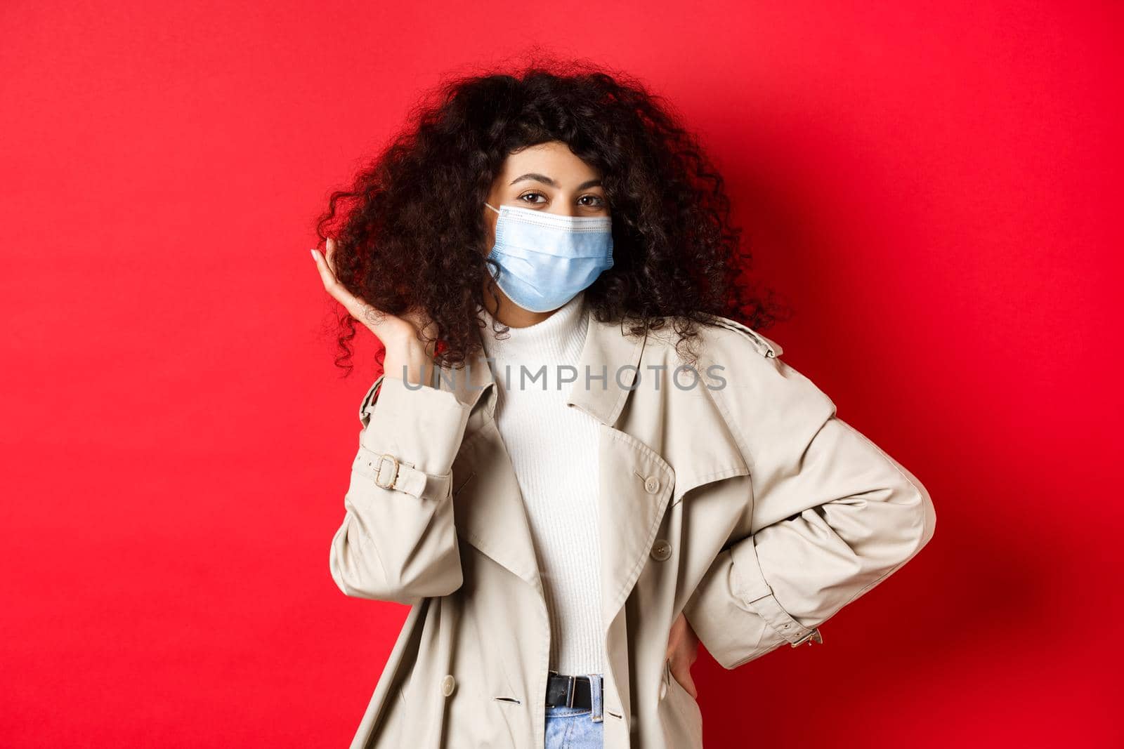 Covid-19, pandemic and quarantine concept. Stylish coquettish woman in medical mask and trench coat, fixing her curly hairstyle and smiling, red background.