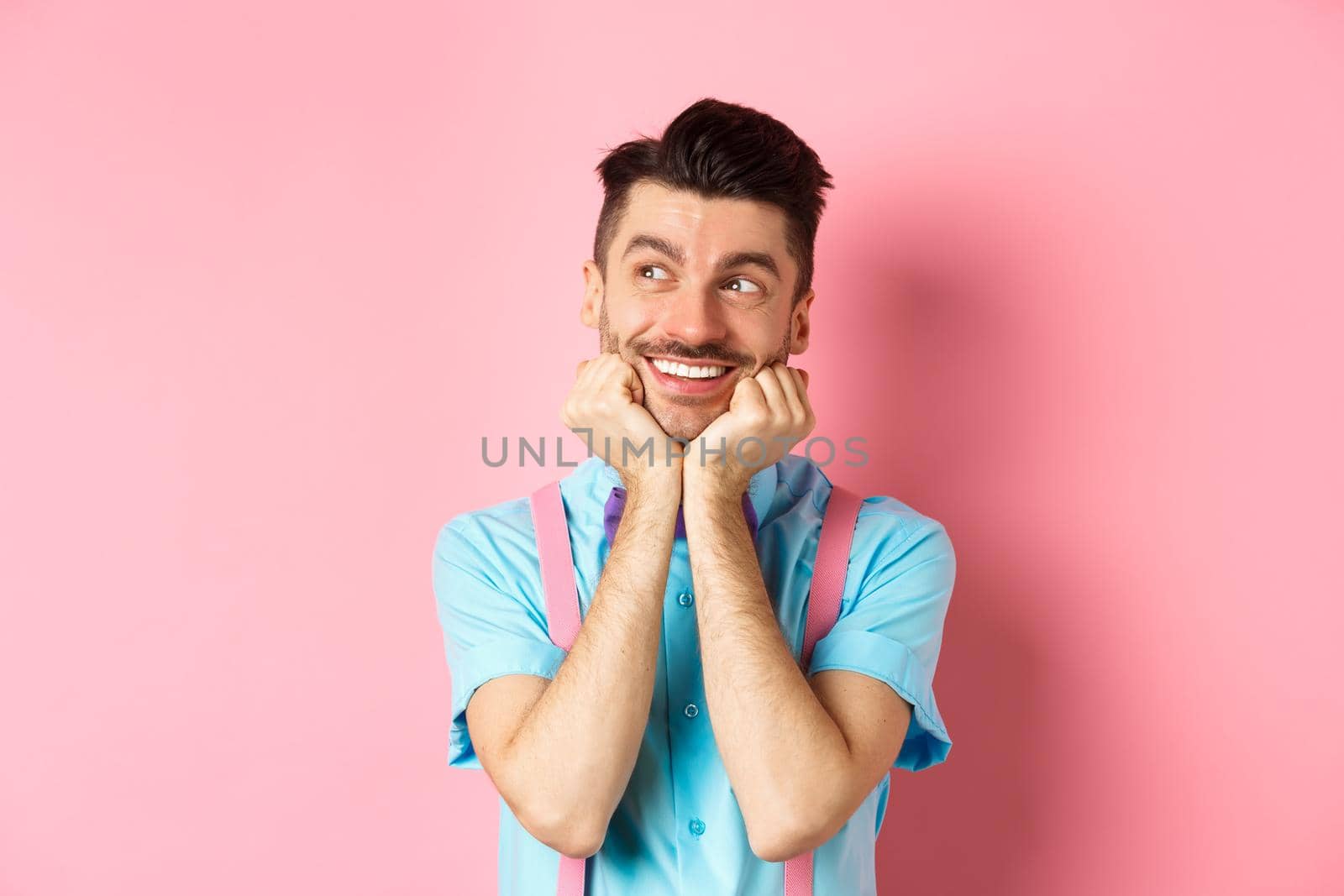 Attractive smiling guy dreaming of something, looking at upper left corner imaging things, daydreaming on pink background. Copy space