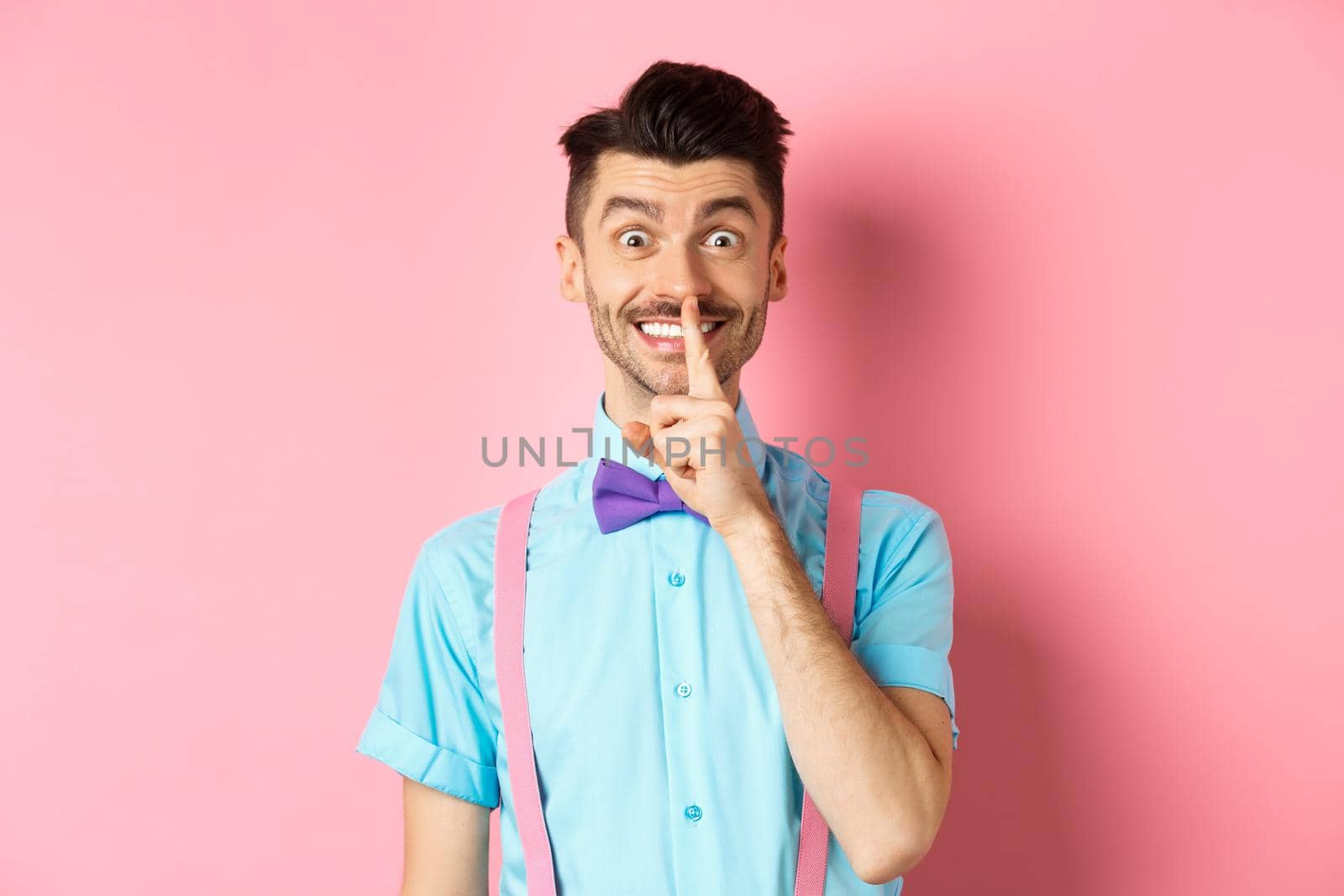 Cheerful young man making surprise, shushing at camera with happy smile, asking to keep voice down, be quiet, standing on pink background by Benzoix