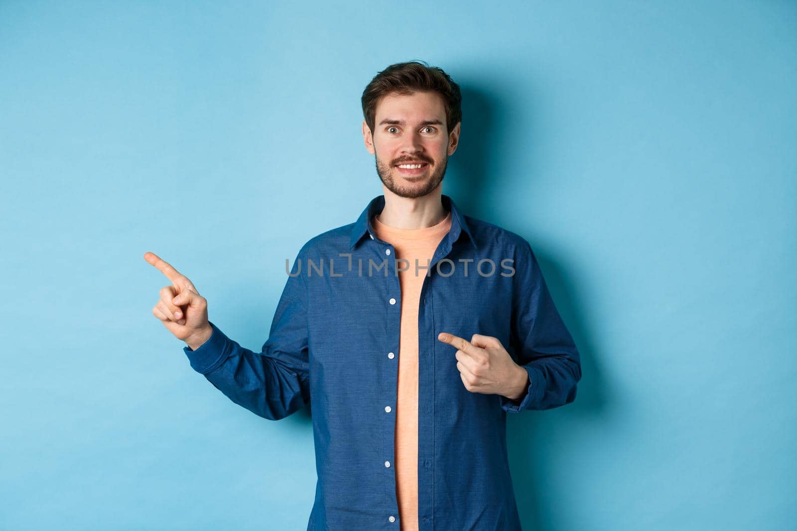 Happy smiling caucasian man showing advertisement, pointing fingers left at empty space, standing on blue background by Benzoix