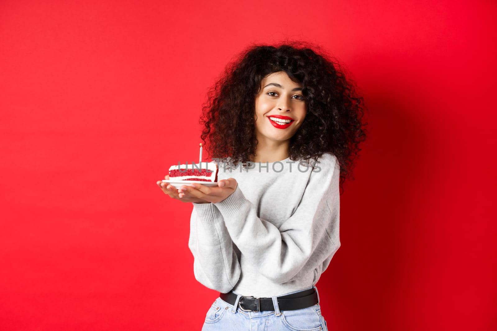 Celebration and holidays concept. Smiling beautiful woman celebrating birthday, holding b-day cake with candle and making wish, standing happy on red background.