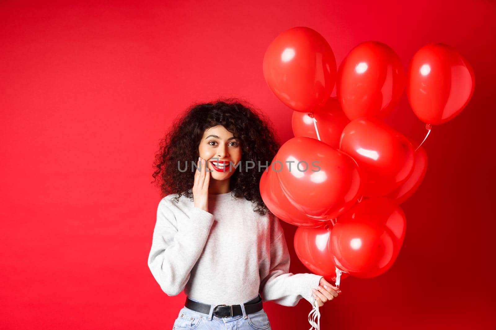 Holidays and celebration. Surprised birthday girl holding helium party balloons and looking touched at camera, being congratulated, red background.