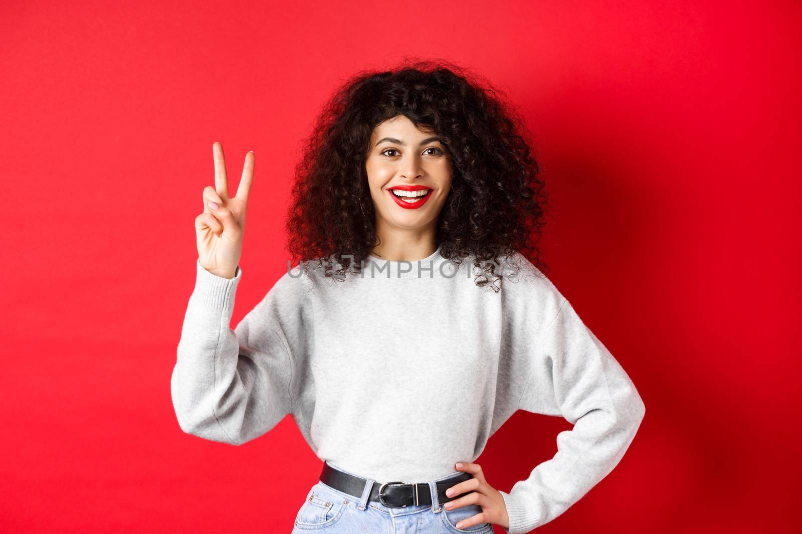 Young pretty lady in sweatshirt showing number two, making an order and smiling, standing on red background.