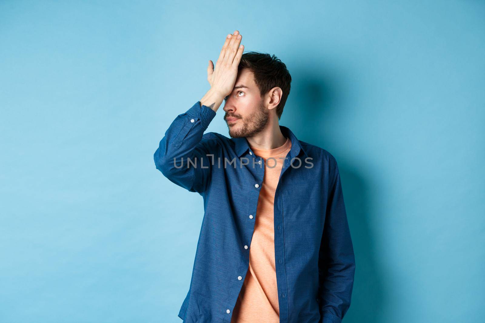 Annoyed and tired man facepalm, roll eyes and look bothered by stupid behaviour, standing on blue background.