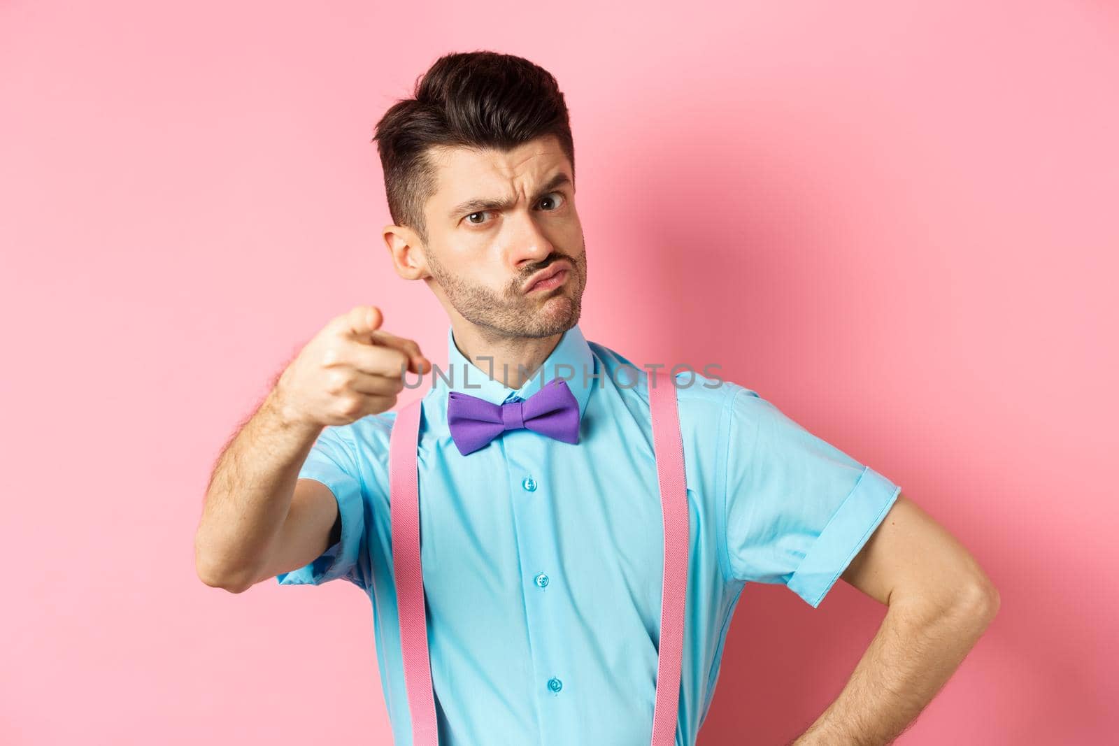 Grumpy guy in bow-tie and suspenders accusing you, its your fault gesture, pointing finger at camera with disappointed frowning face, pink background.