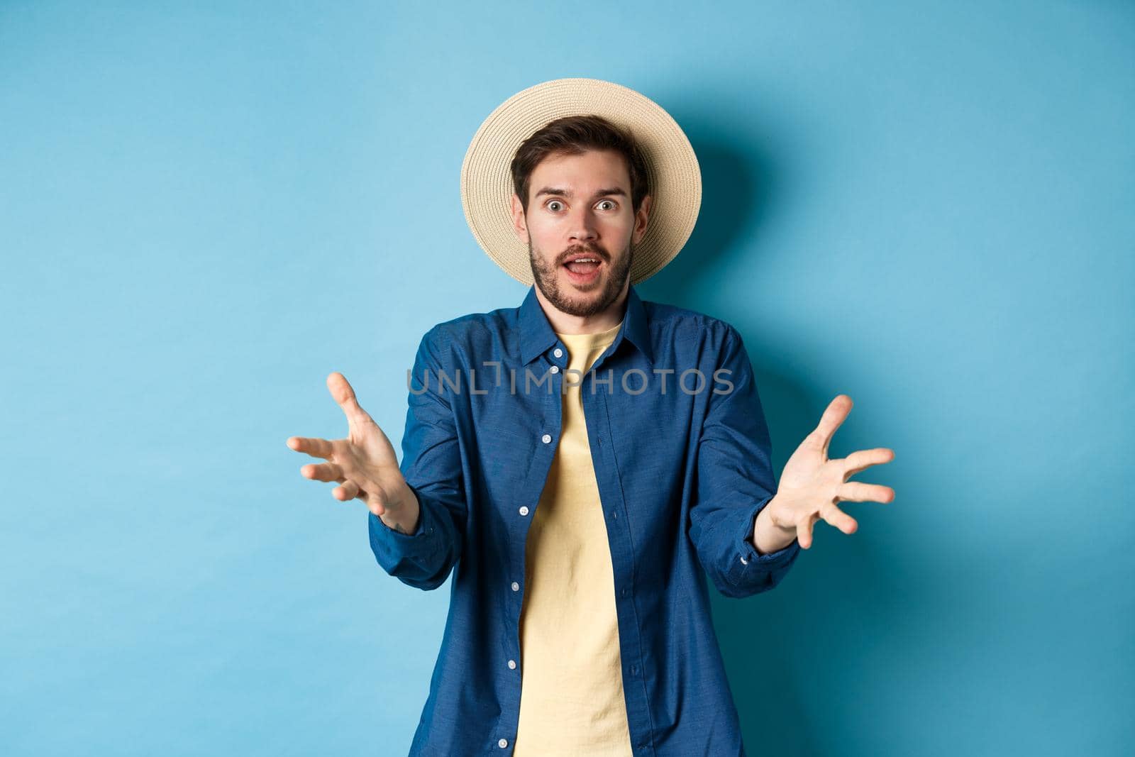 Surprised young man in summer hat, raising hands up staring with disbelief and amazement at camera, impressed with big event, standing on blue background.