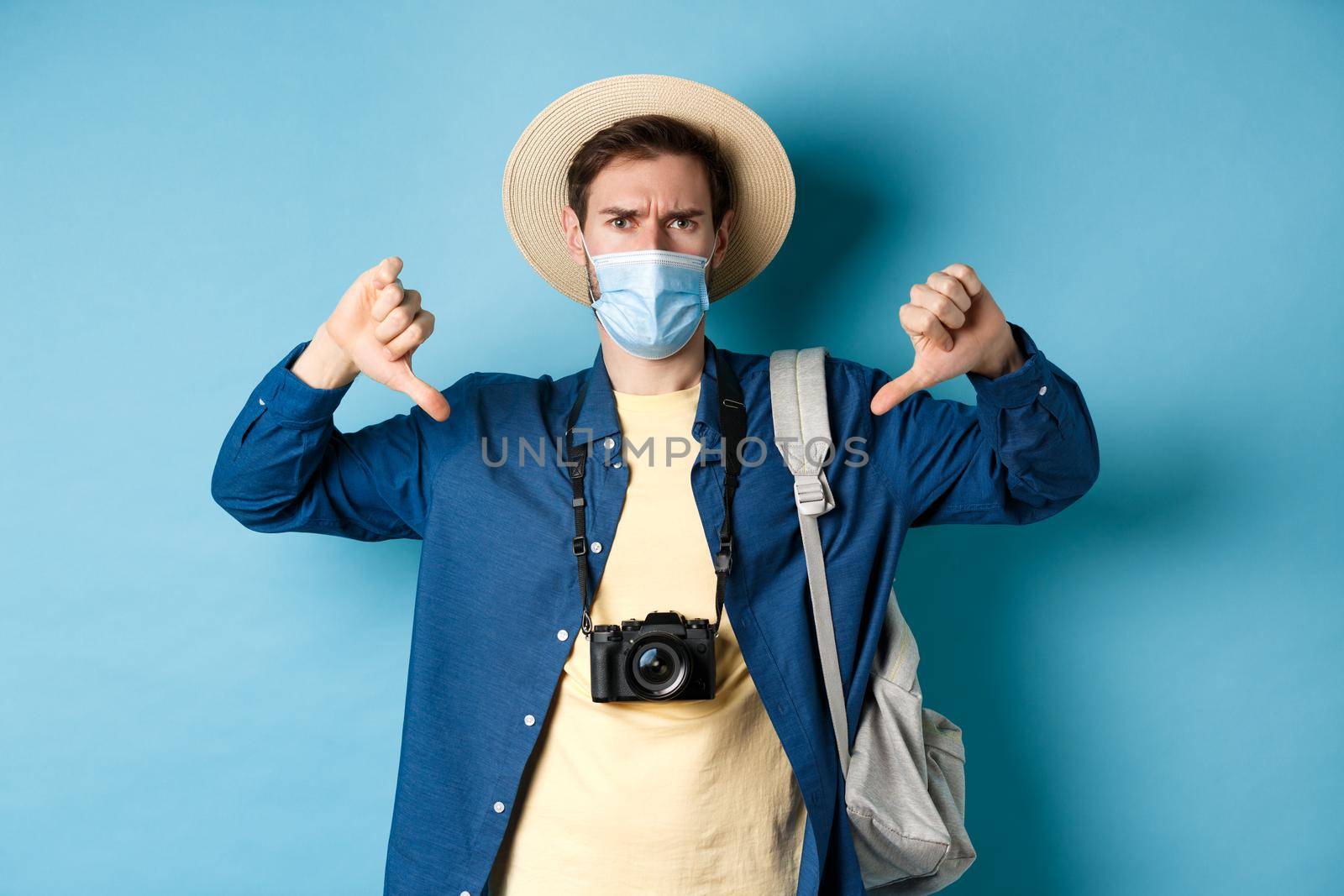 Covid-19 and summer holidays concept. Disappointed tourist in medicla mask frowning, showing thumbs down, negative feedback about travel, standing on blue background.