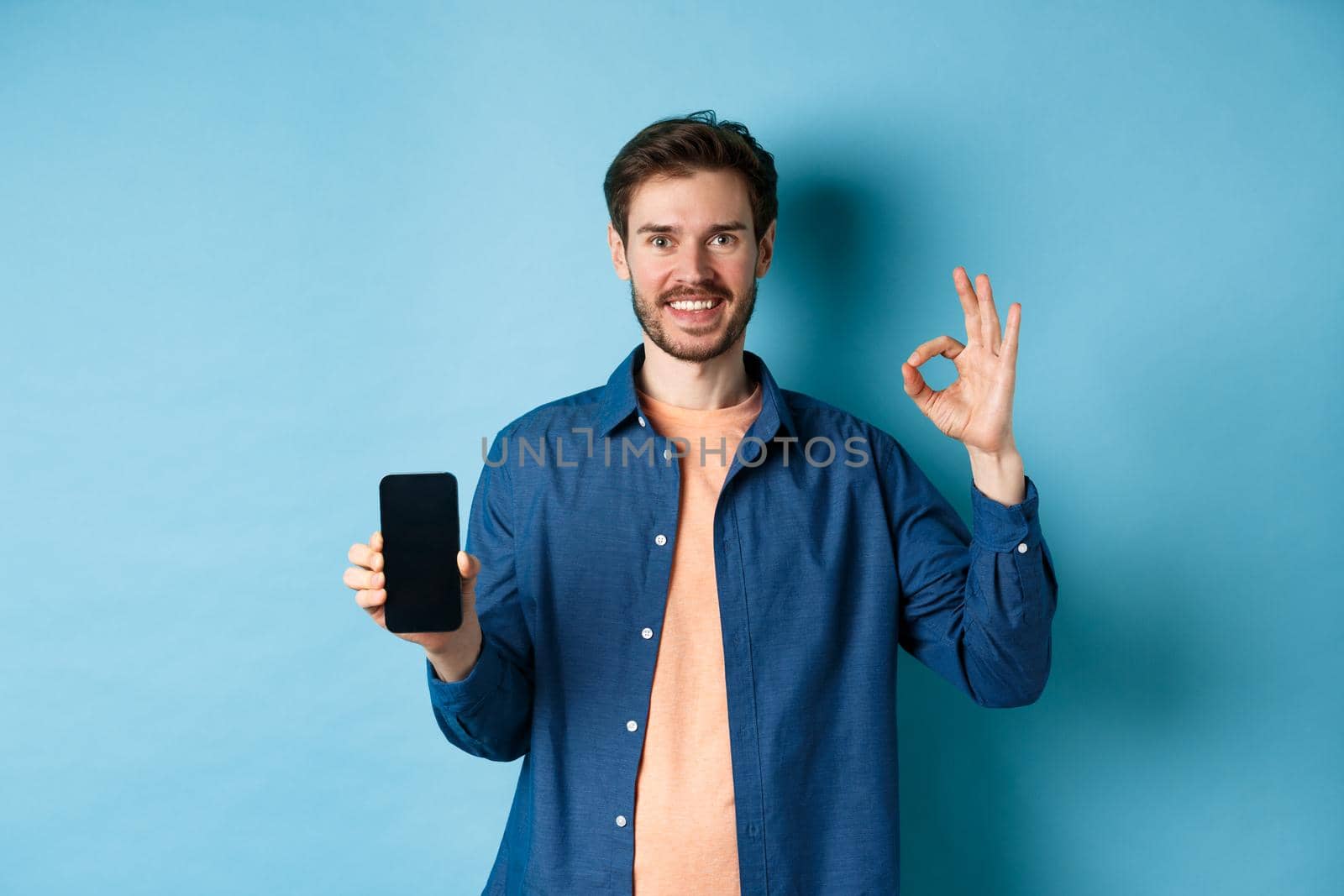 Handsome young man showing okay gesture and empty smartphone screen, recommending app or company, standing on blue background.