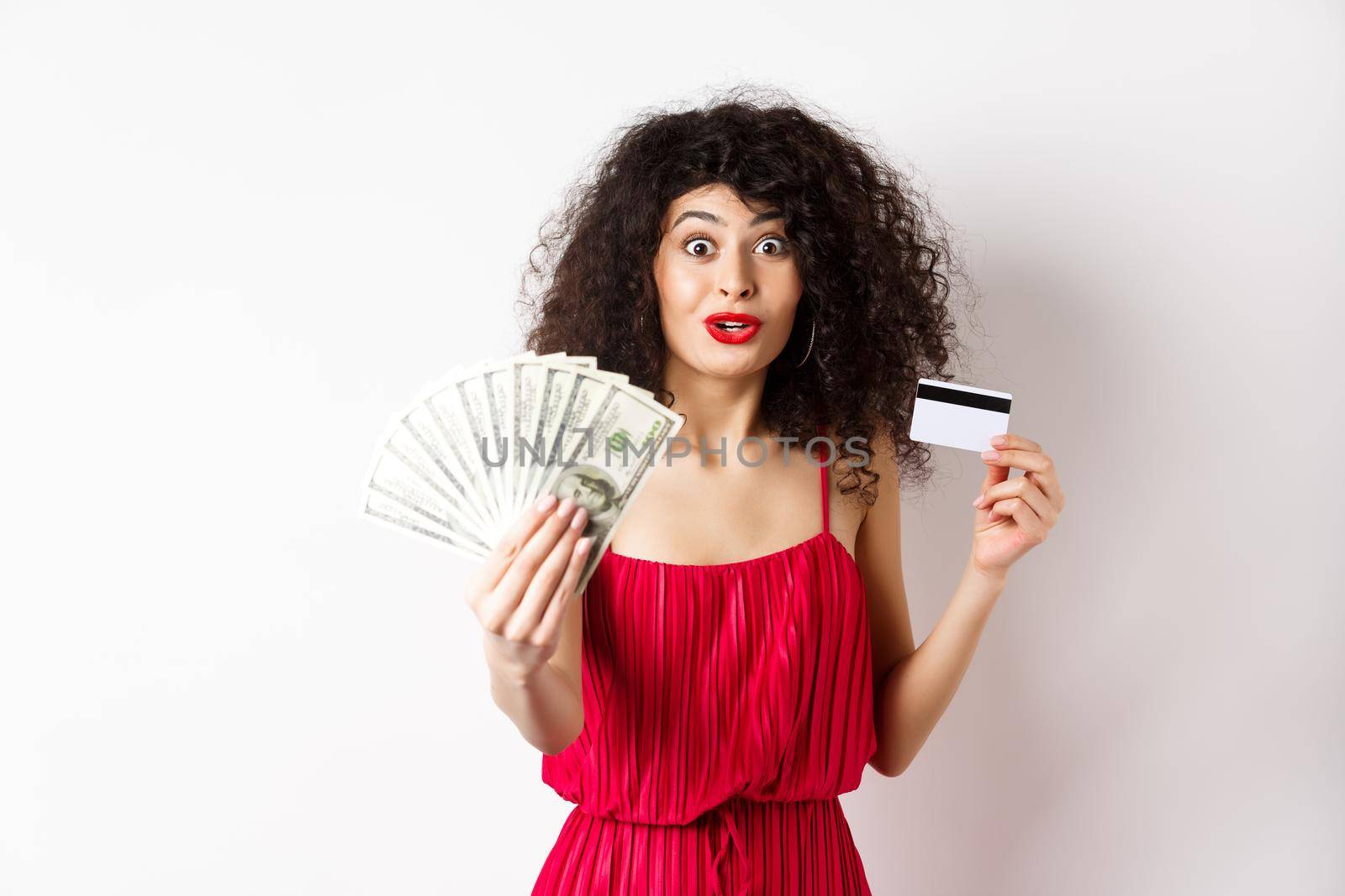 Excited woman looking with amazement and disbelief, showing dollars prize and plastic credit card, standing in red dress on white background.
