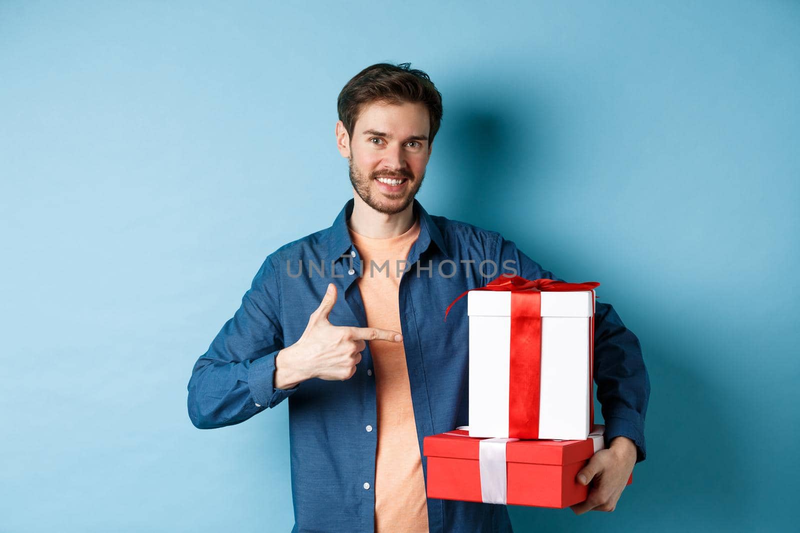 Attractive boyfriend pointing finger at valentines day surprise gifts, showing romantic presents for girlfriend, standing against blue background.