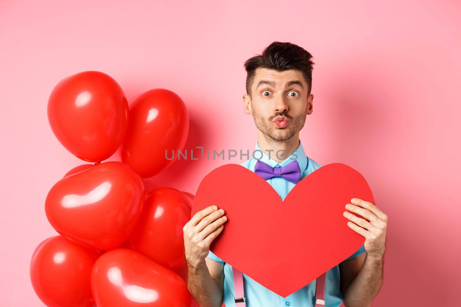 Valentines day concept. Cute guy pucker lips for kiss and showing romantic heart cutout, falling in love, standing over pink background.