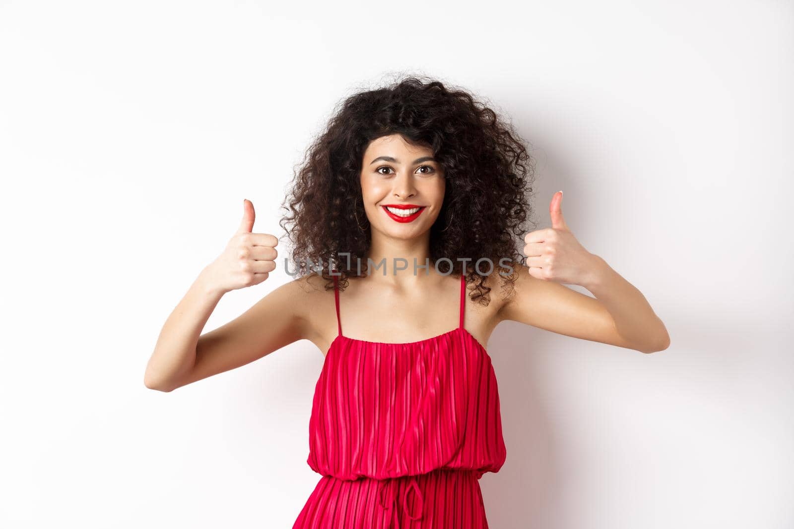 Pretty female model with curly hair and red dress, bright makeup, showing thumbs up in approval, say yes, smiling and nodding in approval, white background.