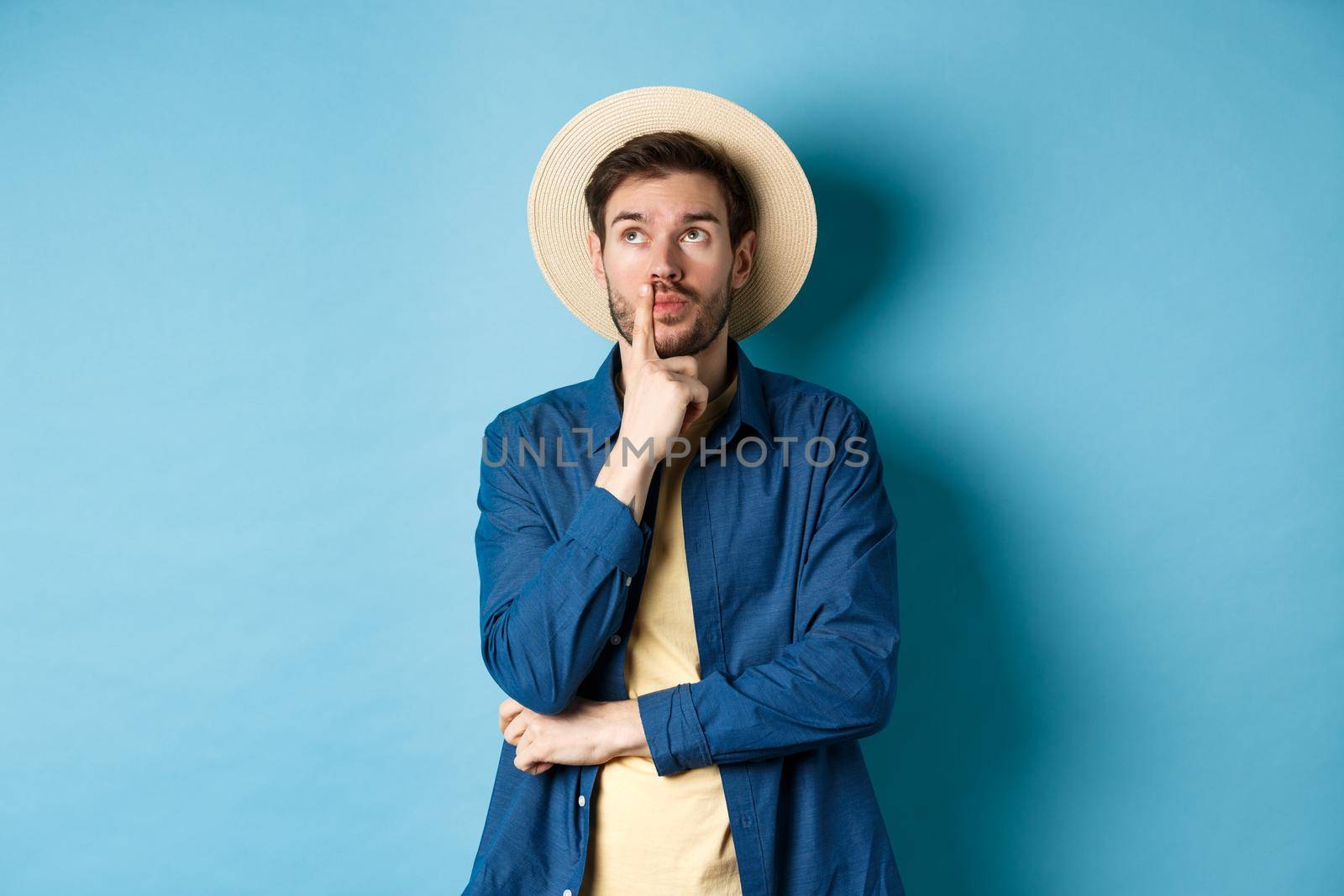 Thoughtful male tourist thinking of summer holiday vacation, looking aside with pensive face, touching lip and pondering next travel, standing on blue background.