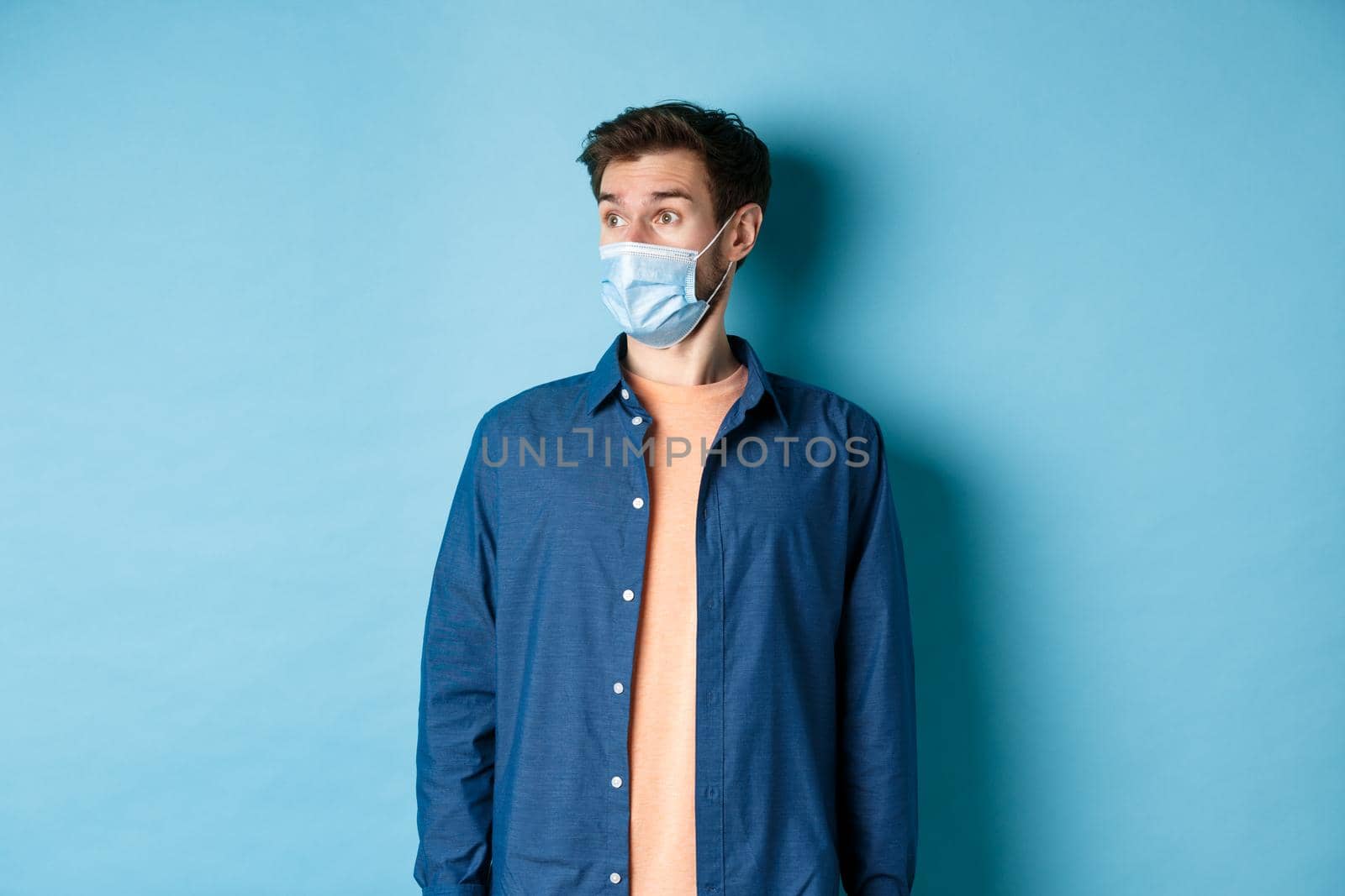 Covid-19 and healthcare concept. Surprised caucasian guy in face mask looking aside at empty space for banner, standing on blue background.