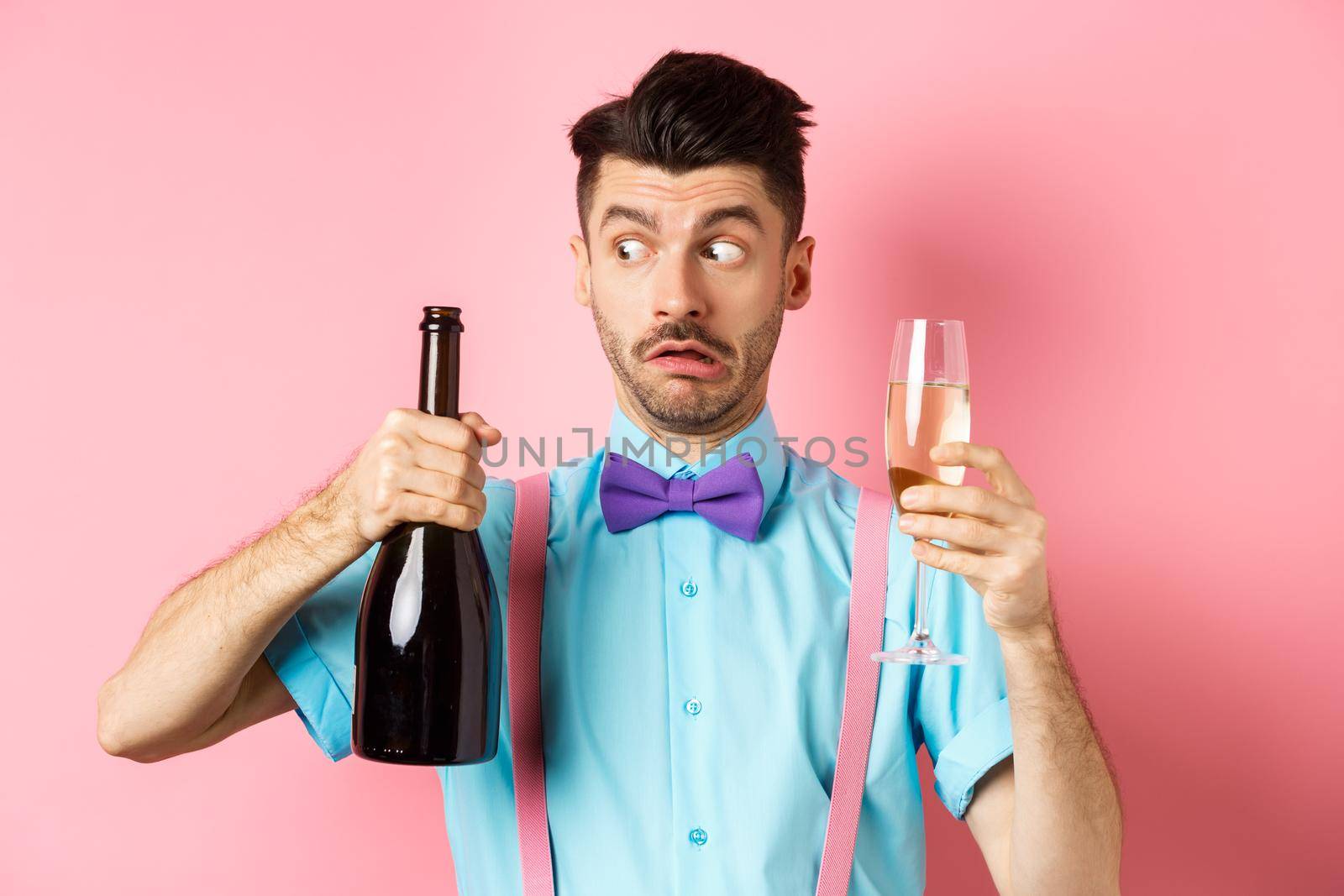 Holidays and celebration concept. Confused drunk guy looking at empty bottle of champagne, holding glass, drinking at party, standing over pink background.