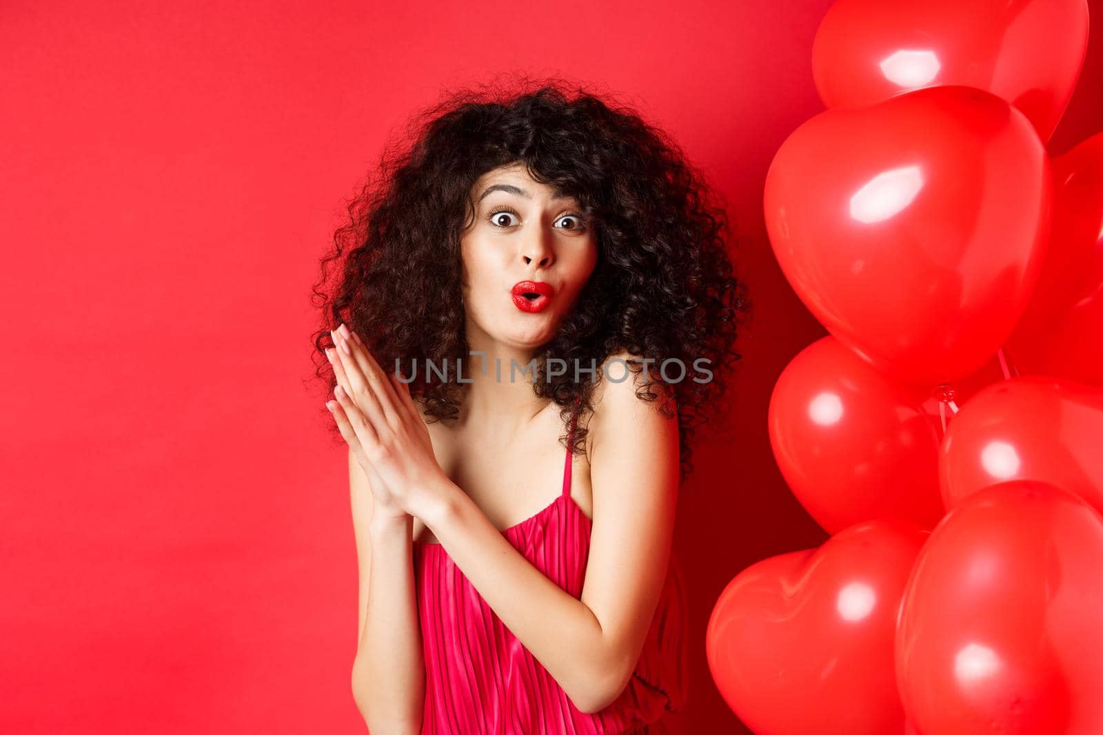 Excited beautiful woman with curly hair, standing near heart balloons and rubbing palms together, expect good deal or relish, red background.