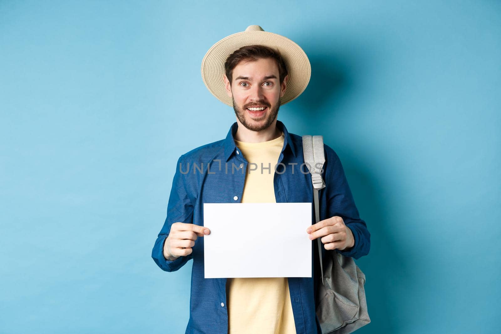 Cheerful tourist in summer hat, showing empty piece of paper and smiling, hitchhiking with backpack, standing on blue background.