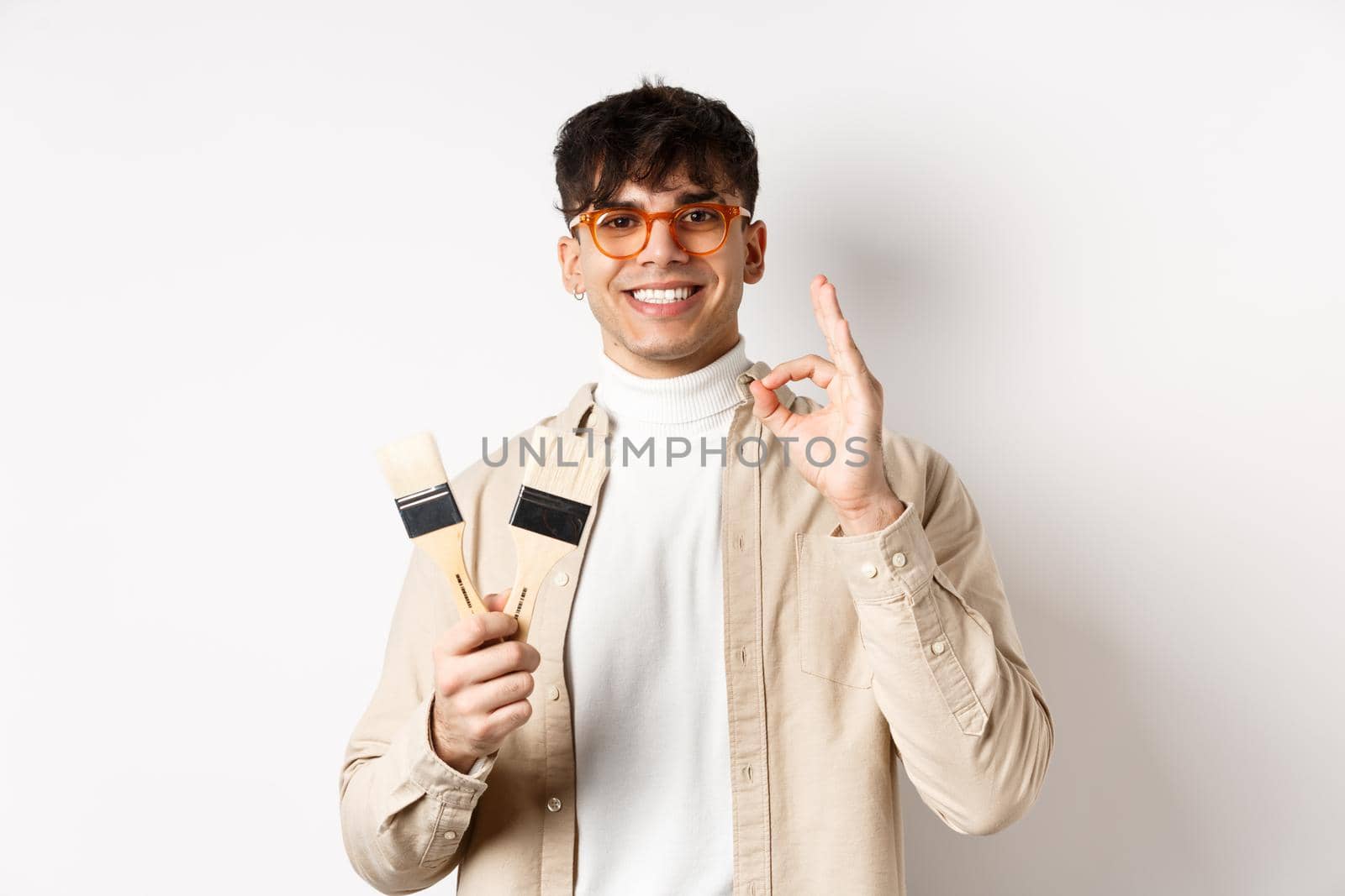 Handsome natural guy in glasses recommending store, showing painting brushes for renovation and decor, make okay sign in approval, standing on white background.