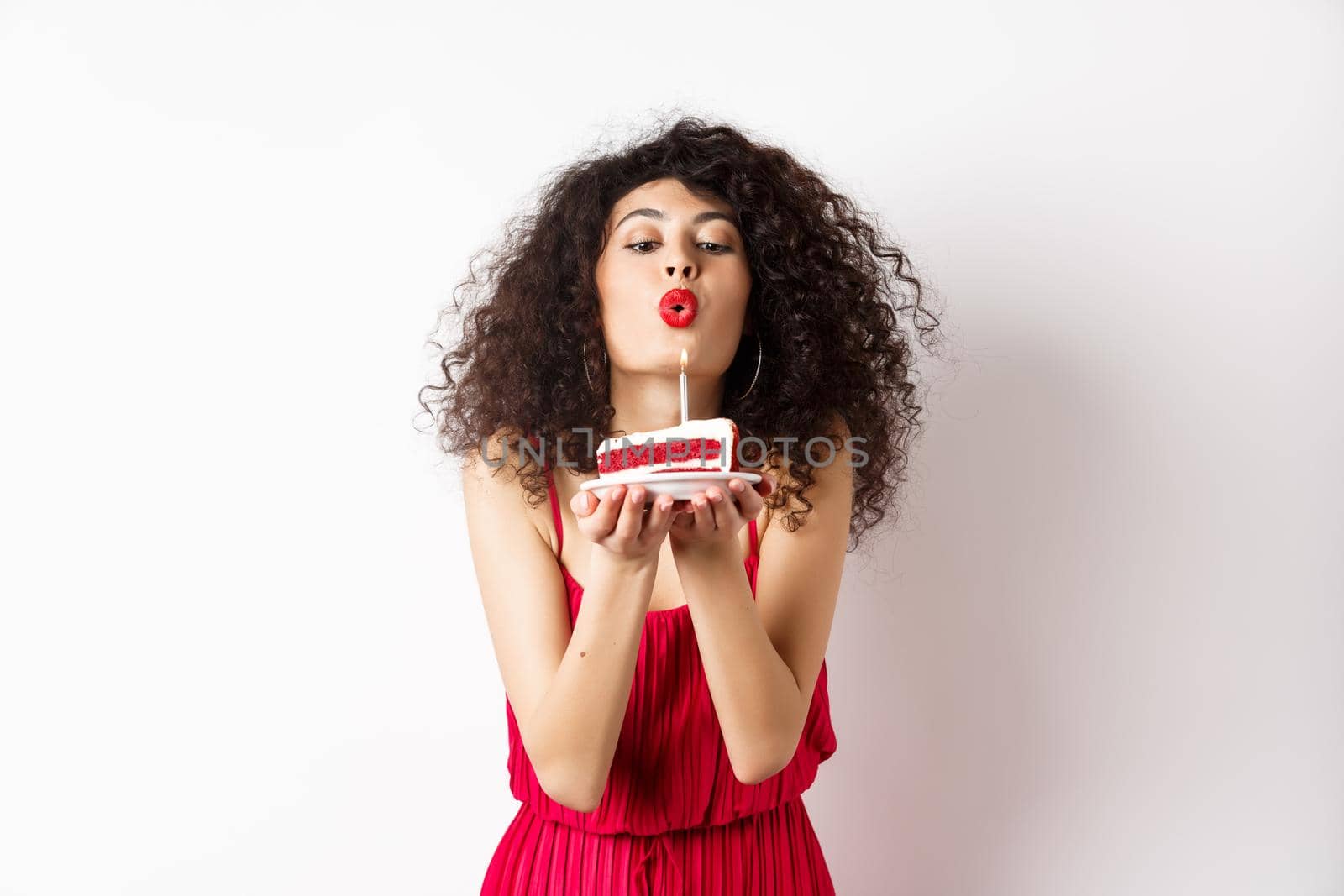 Holidays and celebration concept. Romantic woman in red dress celebrating birthday, blowing out candle on b-day cake and making wish, standing over white background.