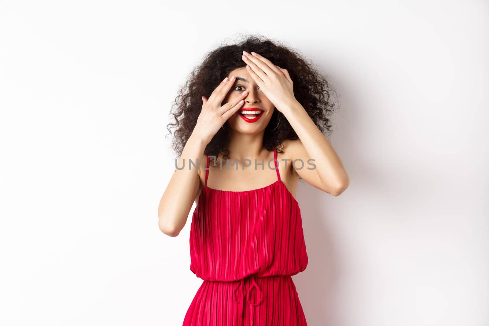 Surprise. Excited pretty girl with curly hair and red lips, peek through fingers and smiling amazed, checking out promo, standing on white background.